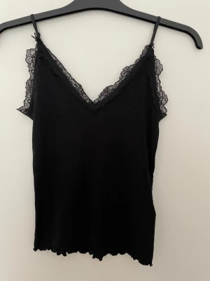 Svart ribbad spetslinne - Säljer ett svart ribbat linne med smala axelband och v-ringad spetskant. Linnet har en figurnära passform och är perfekt för att styla lager på lager. Materialet känns mjukt och stretchigt. Helt och rent men Mindre noppor kan förekomma. 
