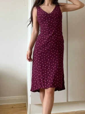 Vinröd prickig klänning från NA-KD - Vinröd ärmlös klänning från NA-KD med rosa prickigt mönster och v-ringning. Klänningen har en figurnära passform och är i midi-längd, perfekt för dig som gillar en feminin och trendig stil. Tillverkad i ett mjukt och lätt material.