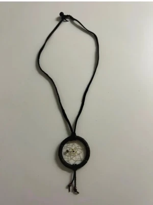 Halsband med drömfångarhänge - Unikt halsband med svart snöre och ett drömfångarhänge i brun ton. Hänget har ett nät av beige tråd och små pärlor i mitten samt längst ner. Perfekt för dig som gillar boho-stil och vill ha något personligt och handgjort.