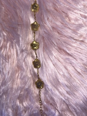 Guldfärgat armband med blommor - Snyggt armband i guldfärg med fem blommor i rad, varje blomma har en liten pärldetalj i mitten. Kedjan är tunn och smidig, perfekt för att ge en elegant touch till din outfit. Materialet ser ut att vara metall med pärldetaljer.