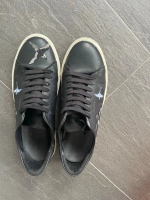 Svarta Artefacto sneakers med fågelmotiv - Svarta sneakers från Axel Arigato med vita sulor och brokiga fågelmotiv i blått, vitt och rött på sidorna och tån. Skorna har svarta skosnören och en klassisk låg profil som passar till många olika stilar. Materialet är slätt läder. Priset kan diskuteras