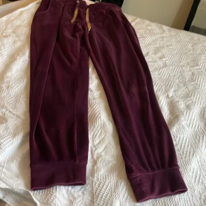 Vinröda mjukisbyxor med gulddetaljer - Säljer ett par vinröda mjukisbyxor med breda muddar och resår i midjan. Byxorna har en snygg dragsko med guldfärgade detaljer och är gjorda i ett mjukt, sammetsliknande material. Perfekta för chill dagar eller när du vill vara extra bekväm.Plysch material.