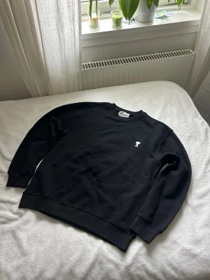Svart sweatshirt från Ami Paris  - Säljer en svart sweatshirt från Ami Paris med rund hals och diskret vit logga på bröstet. Tröjan har en klassisk passform, ribbade muddar vid ärmslut och nederkant samt är tillverkad i mjukt bomullsmaterial.