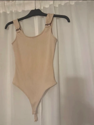 Beige ribbad body - Trendig beige body i ribbat material med breda axelband som knyts i snygga rosetter. Rundad halsringning och tryckknappar i grenen för enkel på- och avtagning. Perfekt att styla till jeans eller kjol för en clean look.