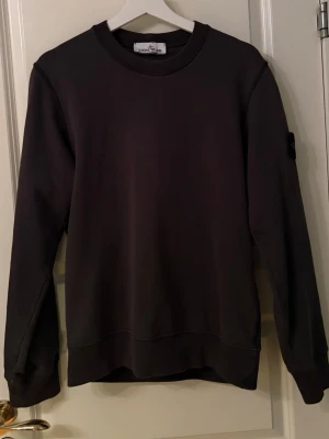 Svart sweatshirt från Stone Island - Säljer en svart sweatshirt från Stone Island med klassisk rund hals och ribbade muddar. Tröjan har den ikoniska Stone Island-loggan på vänster ärm. Perfekt för en clean och stilren look.