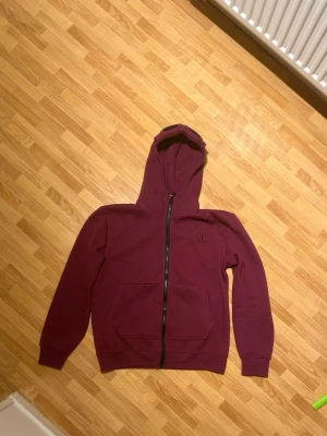 C.P. Company - Snygg vinröd hoodie från C.P. Company med dragkedja framtill och broderad logga på bröstet. Hoodien har en klassisk huva med linsdetalj, samt en stor magficka. Tillverkad i mjuk bomullsblandning och har ribbade muddar vid ärmar och nederkant.