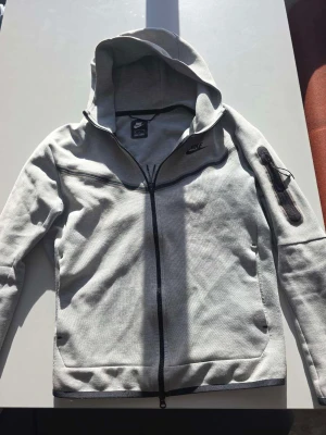 Grå Nike hoodie med dragkedja - Snygg grå hoodie från Nike med hel dragkedja och huva. Har en svart Nike-logga på bröstet och en praktisk ficka med dragkedja på ena ärmen. Tillverkad i mjukt och bekvämt material, perfekt för chill eller träning.