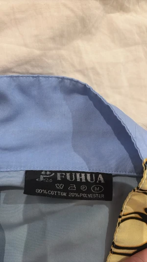 Färgstark skjorta från Fuhua, M - Unik skjorta från Fuhua i storlek M med blå och gul bas, coolt grafiskt mönster med svarta drakliknande figurer och knappar framtill. Tillverkad i 80% bomull och 20% polyester. Perfekt för dig som vill sticka ut med en statement piece.