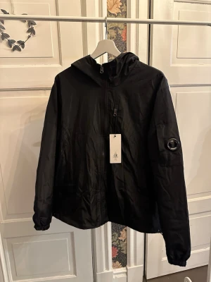 Svart vindjacka från C.P. Company - Snygg svart vindjacka från C.P. Company med huva och dragkedja framtill. Jackan har en ikonisk linsdetalj på ärmen och elastiska muddar. Tillverkad i lätt, vindtätt material som passar perfekt för blåsiga dagar.