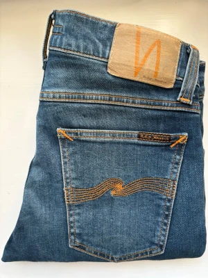 Tight Terry jeans från Nudie Jeans - Snygga blå skinny jeans från Nudie Jeans med klassiska orange sömmar och vågmönster på bakfickan. Modell: Tight Terry, Tvätt: Night Shadow, Waist: 27, Längd: 30. Jeansen har fem fickor, blixtlåsgylf och är tillverkade i mjuk denim. Perfekta för dig som gillar en smal passform och stilren look.