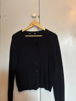 Svart klassisk kofta med rund hals - En enkel och stilren svart kofta med rund hals och knäppning framtill. Koftan har långa ärmar och är tillverkad i ett mjukt material som passar perfekt för lager-på-lager. Snygg att matcha med jeans eller kjol.