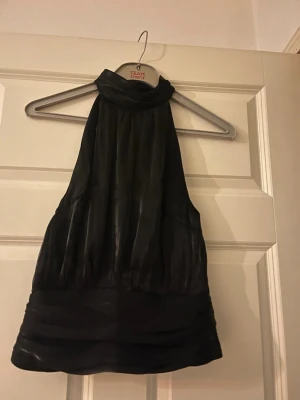 Svart halterneck topp Gina Tricot - Snygg svart halterneck topp från Gina Tricot med hög krage och ärmlös design. Toppen har ett lätt glansigt tyg och bredare band nedtill som ger en cool look. Perfekt att styla med jeans eller kjol för en trendig outfit.