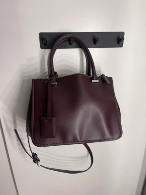 Väska Zara  - Snygg mörklila handväska i slätt skinn med både handtag och justerbar axelrem. Väskan har en stilren, rektangulär form och silverfärgade metalldetaljer. Perfekt för dig som gillar minimalistisk design och vill ha plats för det viktigaste.