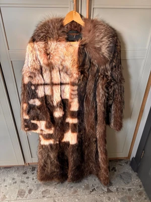 Lång brun pälskappa med bred krage - Säljer en lång, vintageinspirerad pälsrock i olika bruna nyanser. Kappan har en bred, fluffig krage och helsvart innerfoder. Pälsen är tät och ger en lyxig känsla, perfekt för kalla dagar. Stängs framtill och har en klassisk, rak passform. Inköpt på H.Andersson i Göteborg. 