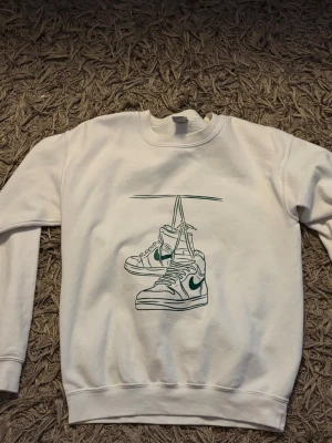 Vit sweatshirt med sneakers-tryck - Vit sweatshirt med ett stilrent tryck av ett par hängande sneakers på bröstet. Tröjan har rund halsringning och ribbade muddar vid ärmslut och nederkant. Perfekt för en avslappnad streetwear-look.