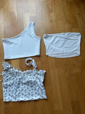 3 vita toppar i olika modeller - Säljer tre olika vita toppar: en oneshoulder ribbad topp, en enkel tubtopp och en blommig linnetopp med volang och knyt fram. Perfekta för sommaren och att mixa och matcha till olika outfits. Säljer på grund av för liten i storlek och brist på andvänding!