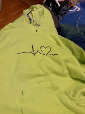 Neongul hoodie med hjärtpuls-tryck - Säljer en neongul hoodie med svart tryck av en hjärtpuls och ett hjärta på bröstet. Hoodien har huva med dragsko och en stor magficka framtill. Perfekt för dig som gillar färgstarka plagg och vill sticka ut.