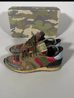 Valentino rockrunner - Snygga sneakers från Valentino med camomönster i grönt, rött och svart. Skorna har detaljer i mocka och textil, samt en ljusblå häl och svarta partier. Klassisk låg modell med gröna skosnören och gummisula. Box med kamouflagemönster medföljer.