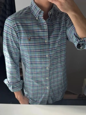 Rutig skjorta från Ralph Lauren - Klassisk rutig skjorta från Ralph Lauren i blå, grön och vit färgskala. Skjortan har lång ärm, knappar framtill och button-down krage. Tillverkad i bomull och har den ikoniska broderade loggan på bröstet. Perfekt för en avslappnad men ändå stilsäker look.