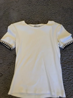 Vit topp med dekorativa ärmslut - Vit topp från Zara med korta ärmar och dekorativa svarta detaljer vid ärmsluten. T-shirten har en rund halsringning och är tillverkad i ett mjukt bomullsmaterial. Perfekt för en stilren och trendig look.