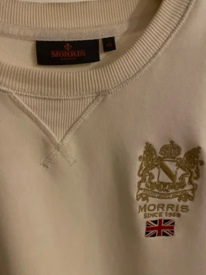 Vit sweatshirt från Morris - Snygg vit sweatshirt från Morris med broderad logga och brittisk flagga på bröstet. Tröjan har rund halsringning, ribbade muddar och klassisk passform. Perfekt för en clean och stilren look.