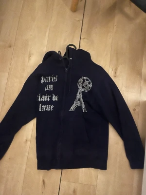 Svart hoodie med Eiffeltorn-tryck - Snygg marinblå hoodie med dragkedja och huva. På framsidan finns ett tryck av Eiffeltornet och texten 'Paris au clair de lune' i vit stil. Hoodien har fickor framtill och justerbar dragsko i huvan. Perfekt för dig som gillar streetwear och Paris-vibbar.