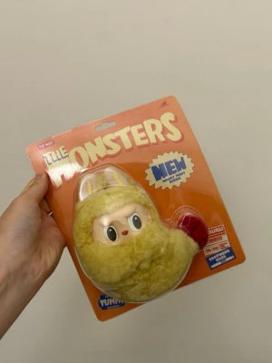 Authentic Labubu Wacky Mart Shrimp Tempura Earphone Case - Säljer en Pop Mart Little Monsters-figur i originalförpackning. Figuren har ett gulligt ansikte med stora ögon och är klädd i en gul fluffig dräkt med öron och ett rött hjärta. Förpackningen är orange och tydligt märkt med serienamnet. Perfekt för samlare eller som dekoration. Hörlurar tillbehör 
