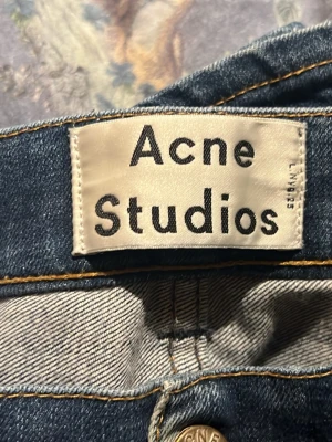 Mörk Blå Acne Studios jeans slim - Säljer ett par klassiska blå jeans från Acne Studios med slim passform. Jeansen har fem fickor, snygga kontrastsömmar och är tillverkade i slitstarkt denim. Perfekta för dig som gillar en tidlös och clean look. finns ett litet hål mellan benen, enkelt att sy igen(tar max 5 min)