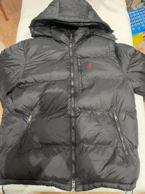 Svart pufferjacka från Polo Ralph Lauren - Snygg svart pufferjacka från Polo Ralph Lauren med huva och två dragkedjeförsedda fickor. Jackan har en röd broderad logga på bröstet och quiltad design. Perfekt för kalla vinterdagar och håller dig varm med sin vadderade insida.