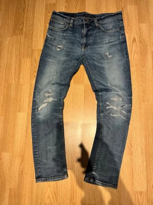 Blå slitna jeans med detaljer - Säljer ett par blå jeans med slitningar och distressed detaljer på både fram- och baksida. Jeansen har klassisk femficksdesign, raka ben och kontrastsömmar. Perfekta för en avslappnad och trendig look.