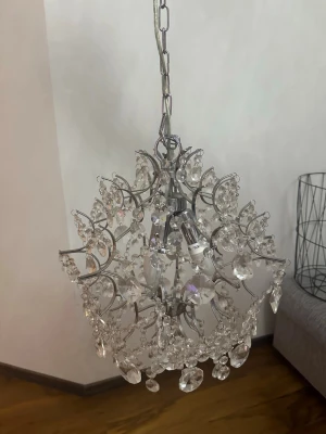 Kristallkrona i silver och glas - Säljer en rund kristallkrona med silverfärgad stomme och hängande glaskristaller. Lampan har flera dekorativa detaljer och plats för tre ljuskällor. Perfekt som statement piece i vardagsrum eller hall.