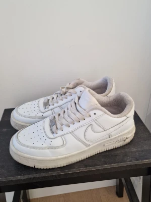 Vita Nike Air Force 1 sneakers - Klassiska vita Nike Air Force 1 sneakers med lågt skaft och perforerad tåbox. Skorna har en tydlig swoosh-logga på sidan, vita skosnören och en robust sula med Air-dämpning. Tillverkade i läder med textilfoder för extra komfort.