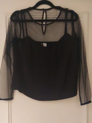 Svart mesh topp med prickigt mönster - Trendig svart mesh topp med transparenta långärmar och små prickar. Toppen har en croppad passform och knyt i nacken. Perfekt för dig som vill ha en edgy och stilren look. Passar till både jeans och kjol.