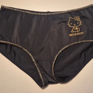Svarta Hello Kitty trosor med glitterkant - Säljer ett par svarta trosor i storlek small från Hello Kitty med guldfärgat tryck fram och bak. Trosorna har glittriga kanter och en liten rosett bak. Materialet är mjukt och stretchigt, perfekt för dig som gillar både söt och lekfull stil. Kommer från ett restlager och är helt oanvända. Se även mina andra anonnser, du får 20% bundlerabatt om du köper flera. 