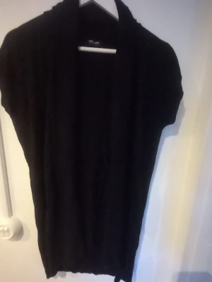 Svart glittrig kortärmad kofta Taifun - Stilren svart glittrig kofta från Taifun Collection med korta ärmar och öppen front. Perfekt att slänga över en topp för en enkel men snygg look. Materialet ser ut att vara mjukt och lättstickat.