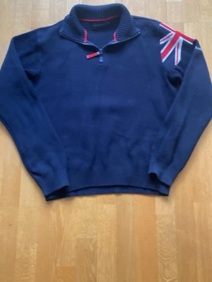 Mörkblå stickad tröja med dragkedja - Mörkblå stickad tröja i 100% bomull med halv dragkedja och hög krage. Snygg detalj med Union Jack-flagga i rött, vitt och blått på ena axeln. Ribbstickade muddar vid ärmslut och nederkant. Perfekt för dig som gillar klassisk men sportig stil. Pris går att diskutera. 
