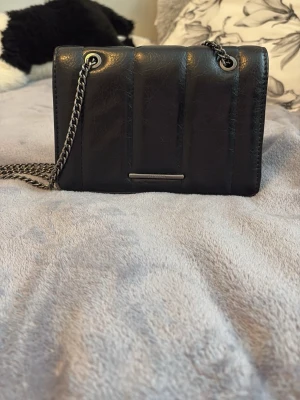 Svart axelväska med kedjerem - Snygg svart axelväska i quiltad look med breda vertikala sömmar. Väskan har en silvrig kedjerem och metallplatta framtill. Perfekt storlek för det viktigaste och ger en cool vibe till din outfit. Jättesnygg passa på att köp!! Inga defekter🫶🏼 skriv om frågor 😋💘