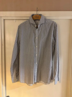 Mönstrad skjorta från bläck - Snygg vit skjorta med blått geometriskt mönster och klassisk krage från Bläck. Skjortan har långa ärmar och knäppning framtill. Perfekt för dig som gillar stilrena detaljer och en fräsch look.