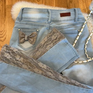 Ljusblå jeans med spetsdetaljer - Unika ljusblå jeans från My Brand med breda spetsinslag längs benen och en söt spetsrosett på bakfickan. Jeansen har hög midja, fem fickor och dekorativa knappar framtill. Perfekta för dig som vill sticka ut med coola detaljer. Sytt bootcut