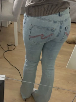 Ljusblå bootcut jeans från Nelly  - Säljer ett par ljusblå bootcut jeans med slitningar vid knäna och rosa detaljer på bakfickorna. Jeansen har klassisk femficksdesign och är tillverkade i denim med lite stretch för skön passform. Perfekta för en avslappnad och trendig look.