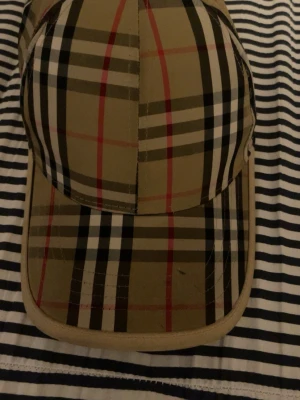 Burberry keps - Snygg beige keps med klassiskt rutigt mönster i svart, vitt och rött. Kepsen har böjd skärm och justerbar rem bak för perfekt passform. Materialet känns som bomull eller polyester och designen är inspirerad av ikoniska brittiska mönster.