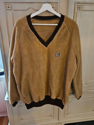 Beige vintage fleece/teddy/fiberpäls tröja Helly Hansen - Mysig vintage beige fiberpäls tröja från Helly Hansen med bruna muddar och V-ringning. Tröjan har en broderad HH-logga på bröstet och är tillverkad i mjukt teddy-material. Perfekt för kalla dagar och enkel att matcha med jeans eller mjukisbyxor.