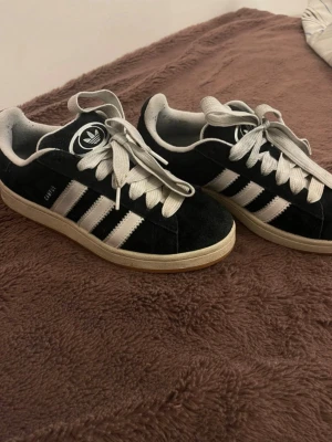 Adidas campus svarta sneakers - Klassiska Adidas campus sneakers i svart mocka med vita detaljer och de ikoniska tre ränderna på sidan. Skorna har vit sula och vita skosnören samt Adidas-logga på plösen. Perfekta för en avslappnad och sportig stil.
