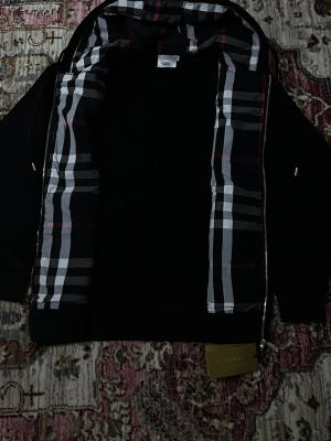 Svart hoodie från Burberry med dragkedja - Snygg svart zipper från Burberry med klassiskt rutigt foder i huvan och längs insidan. Zippern har dragkedja framtill, justerbar huva med snören och ribbade muddar. Perfekt för dig som vill ha en stilren och exklusiv look. Materialet känns mjukt och bekvämt.