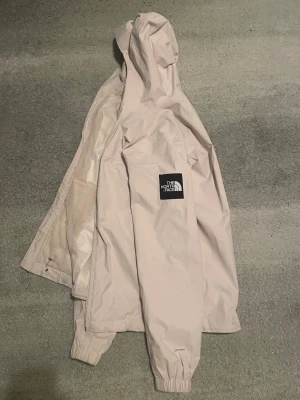 Ljusbeige regnjacka från The North Face - Snygg ljusbeige regnjacka från The North Face med huva och dragkedja. Jackan har elastiska muddar vid ärmsluten, meshfoder på insidan och logga på både bröstet och ärmen. Perfekt för regniga dagar och enkel att matcha med olika outfits.