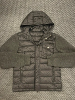 Mörkgrå pufferjacka från baldessarini med stickade ärmar - Snygg mörkgrå pufferjacka med huva och stickade ärmar i matchande grått. Jackan har en bröstficka med knapp, två dragkedjefickor framtill och quiltad design. Perfekt för kalla dagar och ger en cool, avslappnad look.