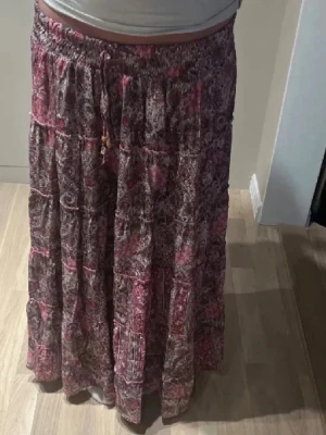 Långkjol från designbysi - Långkjol med blommigt mönster i rosa och lila toner. Den är använd fåtal gånger och i nyskick! Från designbysi i storlek XS-S