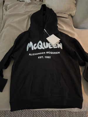 Alexander McQueen hoodie - Säljer en svart hoodie från Alexander McQueen med stor vit logga och texten 'EST. 1992' på bröstet. Hoodien har huva med dragsko och en stor magficka. Materialet känns mjukt och skönt, perfekt för en avslappnad och cool stil. Helt ny passar M och L 