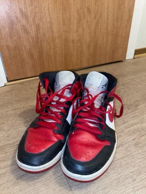 Nike Air Jordan 1 Mid röd/svart/vit - Säljer ett par Nike Air Jordan 1 Mid sneakers i klassisk röd, svart och vit färg. Skorna har läderöverdel, röda snören och ikoniska Swoosh-loggan på sidan. Perfekta för dig som gillar streetwear och vill sticka ut med en tidlös modell.