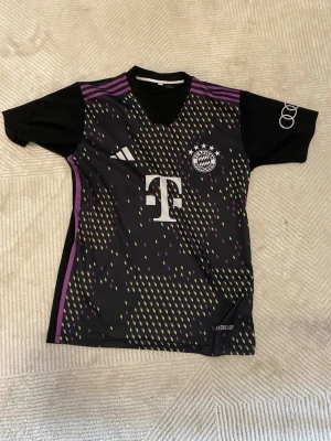 Bayern München Adidas matchtröja svart - Säljer en svart Bayern München matchtröja från Adidas med lila och gula detaljer, vita loggor och klubbmärke på bröstet. Tröjan har korta ärmar, rund hals och är tillverkad i ett lätt funktionsmaterial med AEROREADY-teknologi.